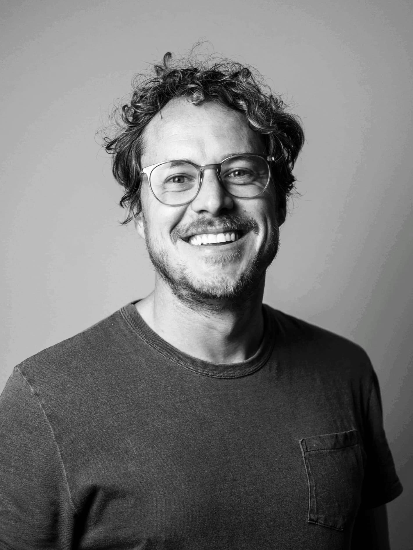 Black and white portrait of Jason Sims smiling, wearing glasses and a t-shirt